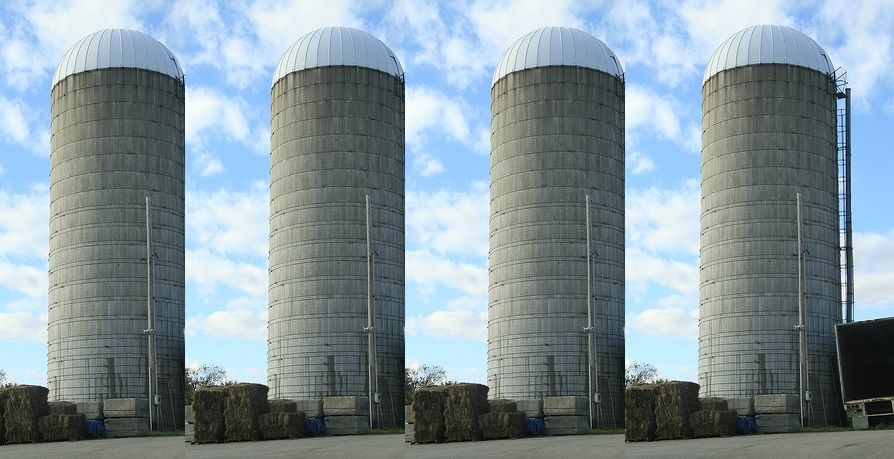 A photograph showing a row of grey silos against a blue sky, with hay bales piled at the foot of each.