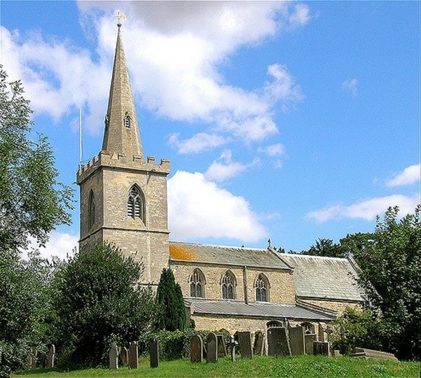 A photograph of a village church with a tower and a spire, underneath a blue sky and surrounded by trees and gravestones.